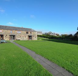 A stone house with wooden doors and windows facing a large lawn with a stone pathway and picnic tables at The Snug in St Minver