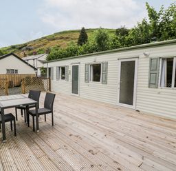 An outdoor wooden deck with a black table and four chairs next to a white caravan with green shutters at Grange Caravan in Llangollen
