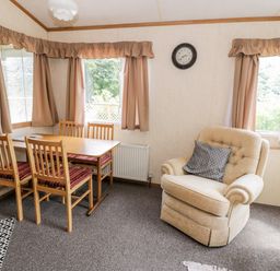 A dining area with wooden table and chairs next to an armchair with a cushion and windows with curtains at Grange Caravan in Llangollen