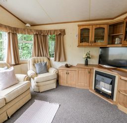 A living room with beige armchairs and a sofa with cushions a carpet in front and a wooden cabinet with a television and fireplace at Grange Caravan in Llangollen