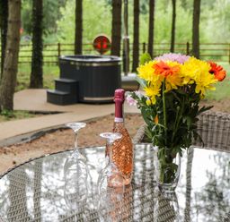 An outdoor glass table with a bottle of rose wine two inverted wine glasses and a vase of yellow orange and pink flowers with a hot tub and trees in the background at Lake View Lodge in Wykeham near East Ayton