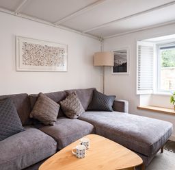 A living room with a grey sectional sofa with pillows a wooden coffee table with two mugs a floor lamp a window with shutters and a small plant on the windowsill at West Cottage in Rock