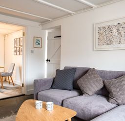 A living room with a grey sofa with cushions and a wooden coffee table with cups next to a dining area with a wooden table and chairs at West Cottage in Rock