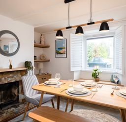 A dining room with a wooden table set for four near a window and a stone fireplace at West Cottage in Rock