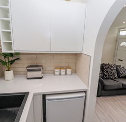A kitchen corner with white cabinets a toaster three labeled jars a plant and a black sink next to a living room with a gray sofa and patterned cushions at 19 Cherry Park in Chapel St Leonards