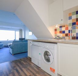 A kitchen area with washing machine dishwasher and colorful tiled backsplash next to a living room with blue sofas and large window at Sea View in Mevagissey