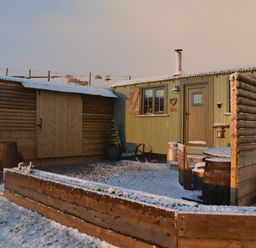 A small wooden cabin with a fenced outdoor seating area and shed in a snowy yard with sheep in the background at Heather Hut at Copy House Hideaway in Earby