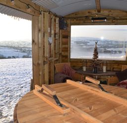 An interior wooden seating area with round table cushions and a window view of snowy hills at Heather Hut at Copy House Hideaway in Earby