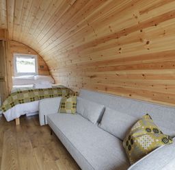 A curved wooden pod interior with a bed, grey sofa, patterned cushions and a bathroom at Dysynni Pod in Llanegryn near Tywyn