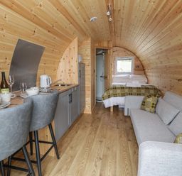 An interior view of a wooden pod with a kitchen area including a sink and kettle two grey bar stools a bed with blankets and pillows a grey sofa with cushions and a small window at Dysynni Pod in Llanegryn near Tywyn