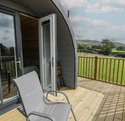 A wooden deck with a chair and open doors leading to a pod with a field and hills in the background at Dysynni Pod in Llanegryn near Tywyn