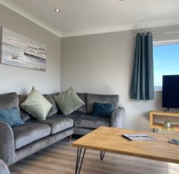 A living room with a grey sofa and blue green cushions a wooden coffee table with magazines a TV on a wooden stand and curtains at Treestumps in Benllech