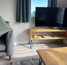A living room with a gray sofa and green cushion a wooden TV stand with a flat screen TV and books and a wooden coffee table at Treestumps in Benllech