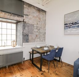 A dining area with a wooden table set for four with blue chairs beside a large window and exposed stone wall at Clarence Riverside Royal William Yard in Plymouth