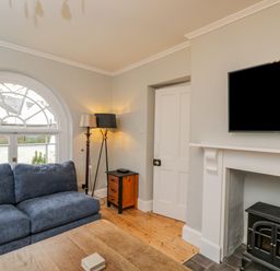 A living room with a blue sofa a wooden coffee table a black stove a television on the wall and a window at White Roses Whitchurch near Monmouth