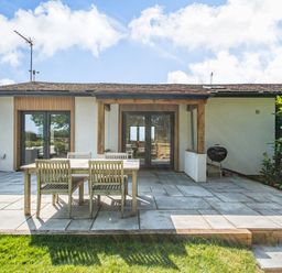 A patio with wooden dining table and chairs in front of a house with glass doors and a barbecue at The Paddock in Honington near Shipston-On-Stour
