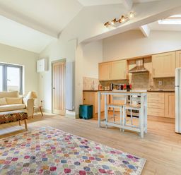 An open-plan living room and kitchen with a sofa, colorful rug, small table with two chairs, wooden cabinets, and a refrigerator at The Paddock in Honington near Shipston-On-Stour