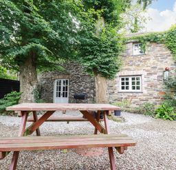 A stone house with a wooden picnic table and trees in an outdoor gravel area at The Cottage in St Kew