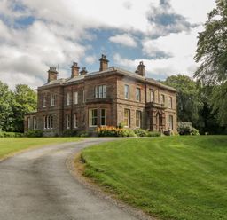 A large brick house with multiple chimneys on a curved driveway surrounded by grass and trees at The Residence in Cleator