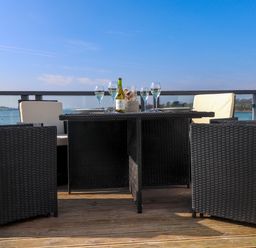 An outdoor dining table with chairs and wine glasses on a wooden deck overlooking water at White Sails in Y Felinheli