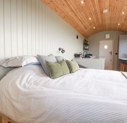 A bedroom with a bed with pillows a wooden ceiling a stove kitchen area and a door at Northcott Hut in Stibb near Bude