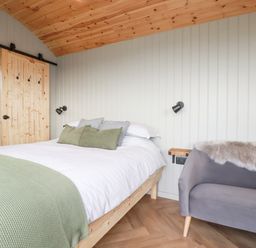 A bedroom with a wooden bed with green pillows and blanket a small gray sofa and a sliding wooden door at Northcott Hut Stibb near Bude