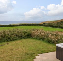A backyard with grassy lawn and bushes with ocean and hills in the background and a hot tub in the corner at Northcott Hut in Stibb near Bude