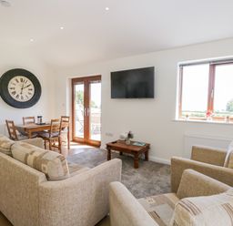 A living room with beige sofas a wooden dining table chairs a wall clock a wall-mounted TV a window and glass doors at Little Barn in Grange-Over-Sands near Cartmel