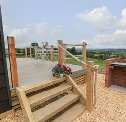 A wooden deck with rope railing, metal chairs and table, flowers, stairs leading down, and a brick barbecue with countryside view at Laird Lodge 1 in Lochmaben