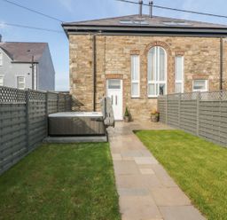 A backyard with a stone pathway leading to a stone house with tall windows and a door fenced by gray wooden panels and a hot tub on the left side at Sardis Chapel 1 Malltraeth