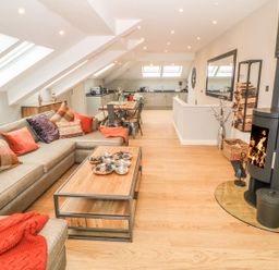 A living room with a large sectional sofa and coffee table facing a wood-burning stove and an open kitchen with a dining table at Sardis Chapel 1 in Malltraeth