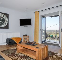 A living room with a wooden coffee table, leather furniture, wall clock, wall-mounted TV, yellow curtains and an open door to a balcony at Escape To The Chapel Anglesey Bryngwran near Rhosneigr