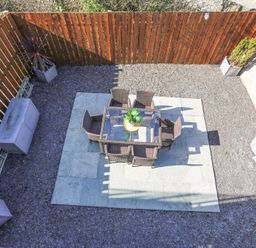 An outdoor patio area with a glass table and six wicker chairs on stone tiles surrounded by gravel and wooden fencing at Escape To The Chapel Anglesey Bryngwran near Rhosneigr