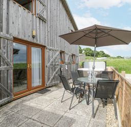 An outdoor patio with a round glass table and four black chairs under a large umbrella and a hot tub beside a wooden house at Top Lodge in Llanvair-Discoed near Penhow