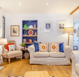 A living room with beige sofas and armchair with cushions bookshelves lamps and framed pictures at Pentreath in Kingsand