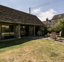An outdoor garden area with lawn wooden bench and table with chairs next to a stone and wood building at Nympy Cottage in Nympsfield near Nailsworth