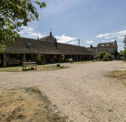 A gravel driveway with a wooden picnic table and benches outside a long single-story stone building with a tiled roof at Nympy Cottage in Nympsfield near Nailsworth