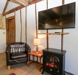 A room with a black armchair a wooden side table with a lamp a black electric fireplace and a wall-mounted tv at Nympy Cottage in Nympsfield near Nailsworth