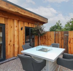 An outdoor patio with a white table with a glass fire feature and four gray chairs next to a wooden building and fence at Daffodil in Llangadfan