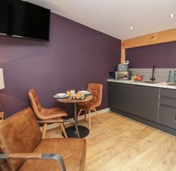 A kitchen area with a small round table and two chairs a brown armchair a wall-mounted TV and kitchen cabinets with appliances at Daffodil in Llangadfan