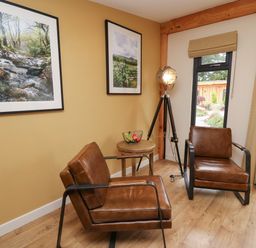 A sitting area with two brown leather chairs a wooden side table with fruit a tripod floor lamp and framed pictures on the wall at Rhosyn in Llangadfan