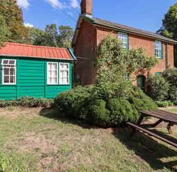 A brick house with green wooden extension and a wooden picnic table in a garden at Warren Cottage in Bures near Assington