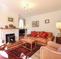 A living room with a fireplace a brown sofa with red cushions two armchairs a wooden coffee table a dark wooden cabinet a lamp and a television at Warren Cottage in Bures near Assington