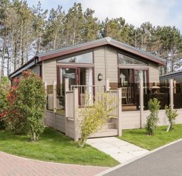 A small beige cabin with large windows and glass railing on a green lawn with shrubs at Rockpool Retreat in Runswick Bay near Hinderwell