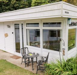 A white exterior corner of a house with large windows a glass door a small round metal table and two chairs a bird feeder and greenery at Ty Haf in Roch