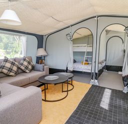 A seating area with a sofa with plaid cushions a floor lamp and two round tables in a tent with bunk beds visible through doorways at The Salmon in Nantmel near Rhayader