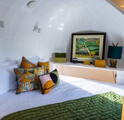 A bed with patterned pillows and a green quilt with artwork and decorative items on a shelf in a white curved room at Veiko - Mayflower Meadow in Burshill near Brandesburton