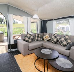 An interior view of a tent with a grey L-shaped sofa with plaid cushions a round black coffee table on a yellow rug and a hot tub outside at The Otter in Nantmel near Rhayader