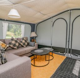 An indoor seating area with a sofa, plaid cushions, two round tables, a floor lamp, and a window with white curtains at The Heron in Nantmel near Rhayader