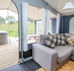 An indoor seating area with a gray couch and plaid pillows next to an outdoor wooden deck with a hot tub and picnic table at The Heron in Nantmel near Rhayader